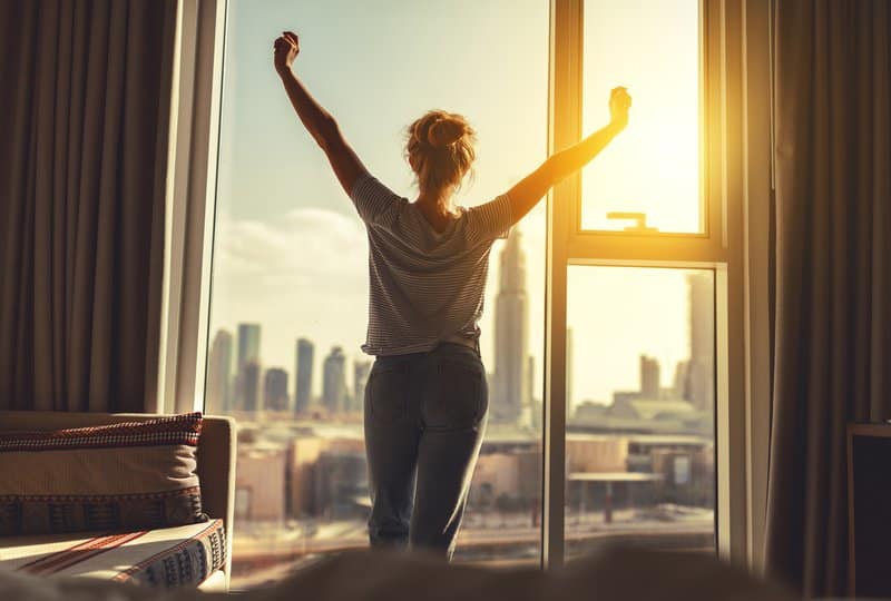 Happy woman stretches and opens curtains at window in morning